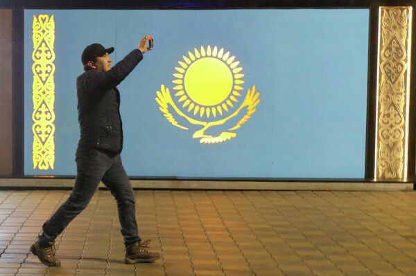 Мужчина держит мобильный телефон, проходя мимо доски с государственным флагом Казахстана во время акции протеста. - Sputnik Азербайджан