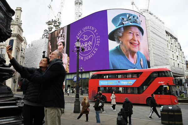 Cütlük 6 fevral 2022-ci ildə Londonun mərkəzində yerləşən Pikadilli meydanında Böyük Britaniya Kraliçası II Elizabetin təsviri ilə rəqəmsal ekran fonunda selfi çəkir. - Sputnik Azərbaycan