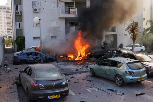 Сотрудник израильских сил безопасности стоит возле горящих автомобилей после ракетного обстрела из сектора Газа в Ашкелоне, на юге Израиля. - Sputnik Азербайджан