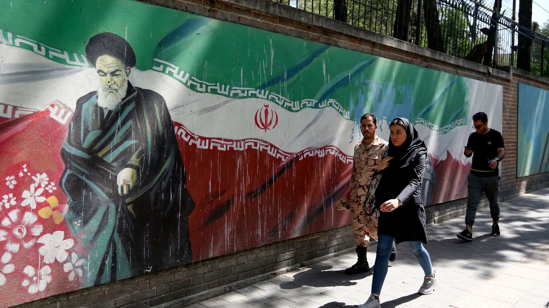 Люди проходят мимо фрески с изображением основателя исламской республики аятоллы Рухоллы Хомейни и государственного флага вдоль стены бывшего посольства США в иранской столице Тегеране 22 июня 2019 года - Sputnik Азербайджан, 1920, 11.02.2022