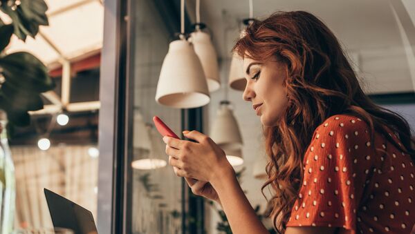 Рыжеволосая девушка в красном платье сидит в кафе и смотрит на свой смартфон - Sputnik Азербайджан