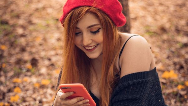 Красивая девушка с длинными рыжими волосами и красном берете смотрит видео на своем смартфоне - Sputnik Азербайджан