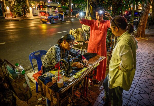 Женщина освещает мобильными телефонами рабочее место уличной швее в Бангкоке, Таиланд  - Sputnik Азербайджан